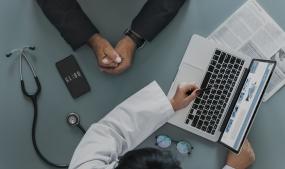 Plan de haut sur un bureau de médicin sur lequel on peut voir un stétoscope, un téléphone, des lunettes puis un ordinateur portable déposé sur un journal. D'un côté du bureau, les bras d'une docteur, avec un stylo à la main gauche, pointant vers l'écran de l'ordinateur. De l'autre côté, les bras d'un patient, mains ensemble.